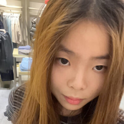 A girl with dyed brown hair wears a striped blue-white crop top and a black side-bag. She stands in front of racks of clothing.