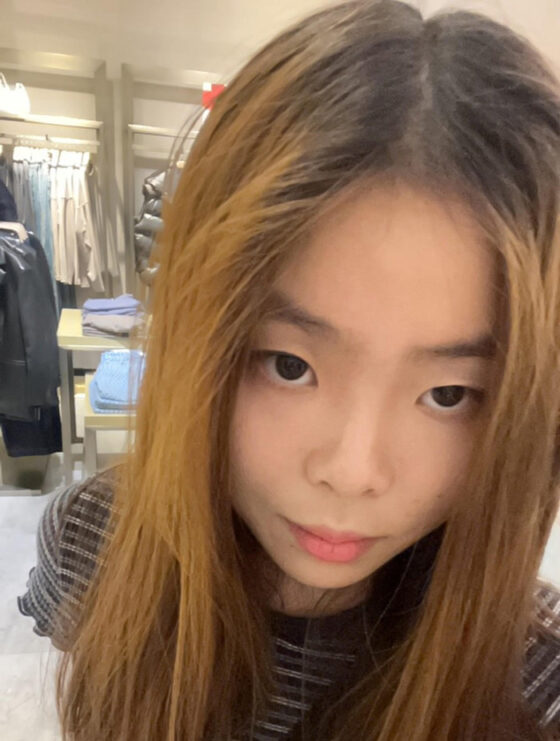 A girl with dyed brown hair wears a striped blue-white crop top and a black side-bag. She stands in front of racks of clothing. 