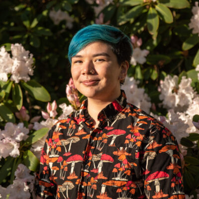 Ally Ang, a gaysian poet with short blue hair, nose ring, and a black shirt printed with bright red and orange mushrooms