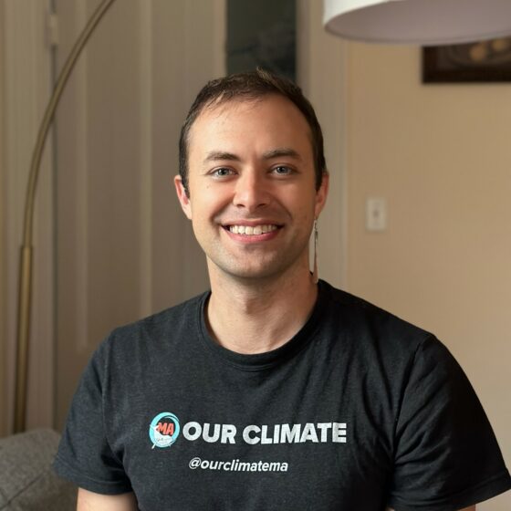 Eben Bein has short hair and smiles into the camera. They wear a black tshirt that says "Our Climate."