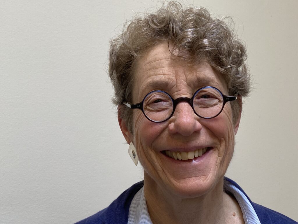 Poet Rebekah Wolman, a white Jewish woman with short curly hair and round-rimmed glasses, smiles widely at the camera