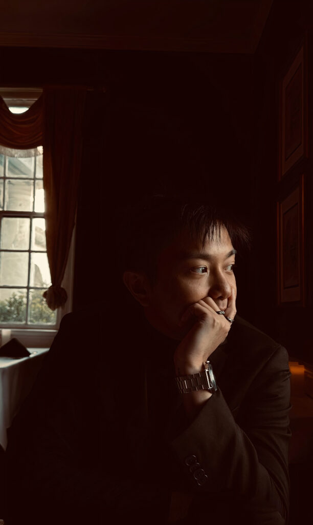 A person in a black blazer sits in a soft window light, their chin resting on their left hand while gazing out the window.