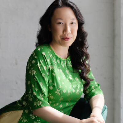 Asian femme seated in a teal-colored velvet chair by a sunny window. She has long black hair in gentle waves and wears a green tunic with golden embellishments over yellow pants.