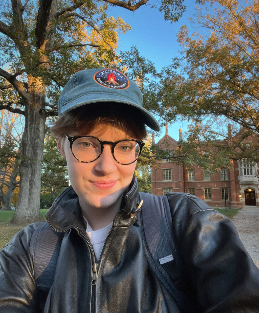 A white, queer person, wearing glasses, a blue hat, a black leather jacket, and a backpack. They are smiling at the camera, with autumnal trees and a blue sky in the background.