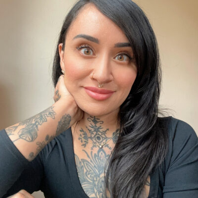 Person wearing a three-quarter sleeve fitting black scoop neck shirt. They have long glossy black hair and tattoos.