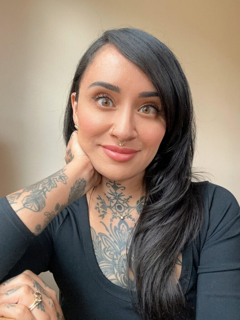 Person wearing a three-quarter sleeve fitting black scoop neck shirt. They have long glossy black hair and tattoos.