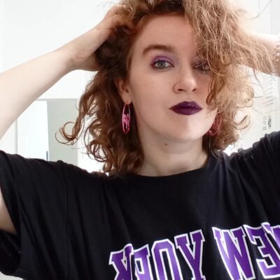 Color photograph of the author, Kathryn Bratt-Pfotenhauer, a white person with shoulder length curly red-blonde hair and green eyes. She wears purple eyeshadow, dark purple lipstick, pink hoop earrings and a partially visible black and purple New York University t-shirt; her hands are in her hair.