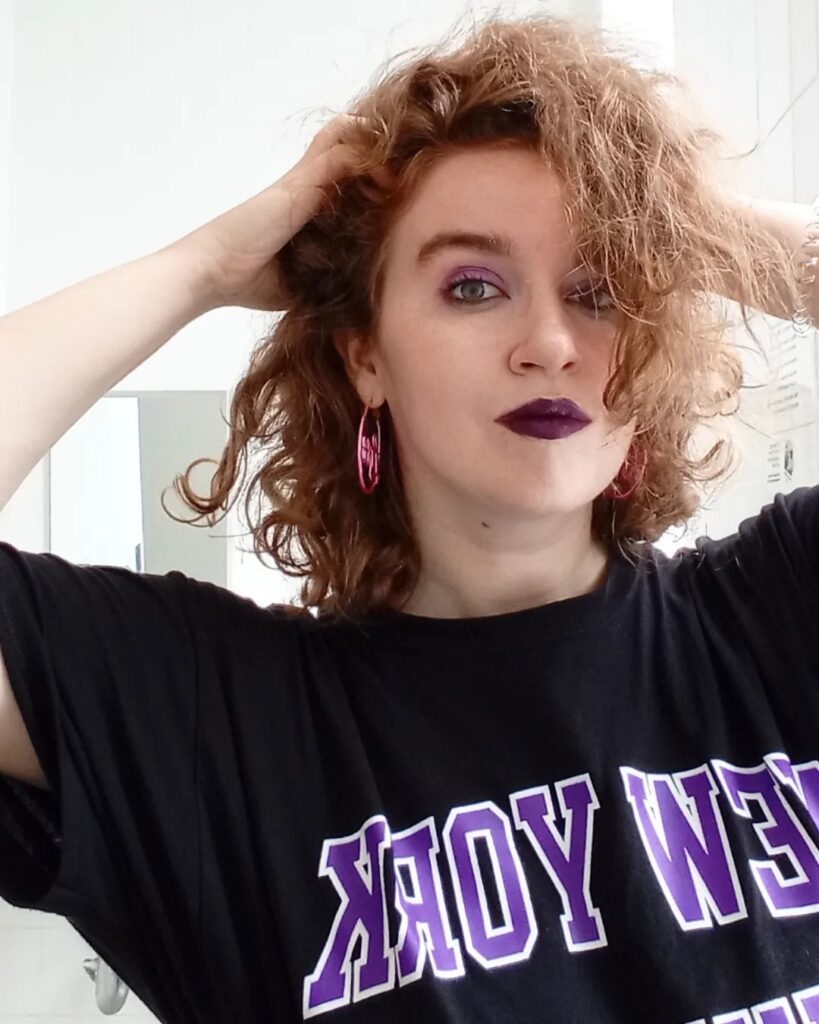 Color photograph of the author, Kathryn Bratt-Pfotenhauer, a white person with shoulder length curly red-blonde hair and green eyes. She wears purple eyeshadow, dark purple lipstick, pink hoop earrings and a partially visible black and purple New York University t-shirt; her hands are in her hair.