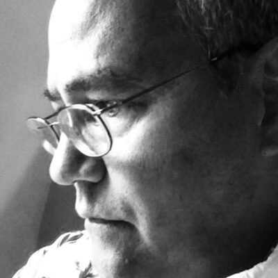 Black and white: A mixed-race white-Japanese man in contemplation by a sunny window. He wears a collared print shirt and a pair of glasses perched on his nose.