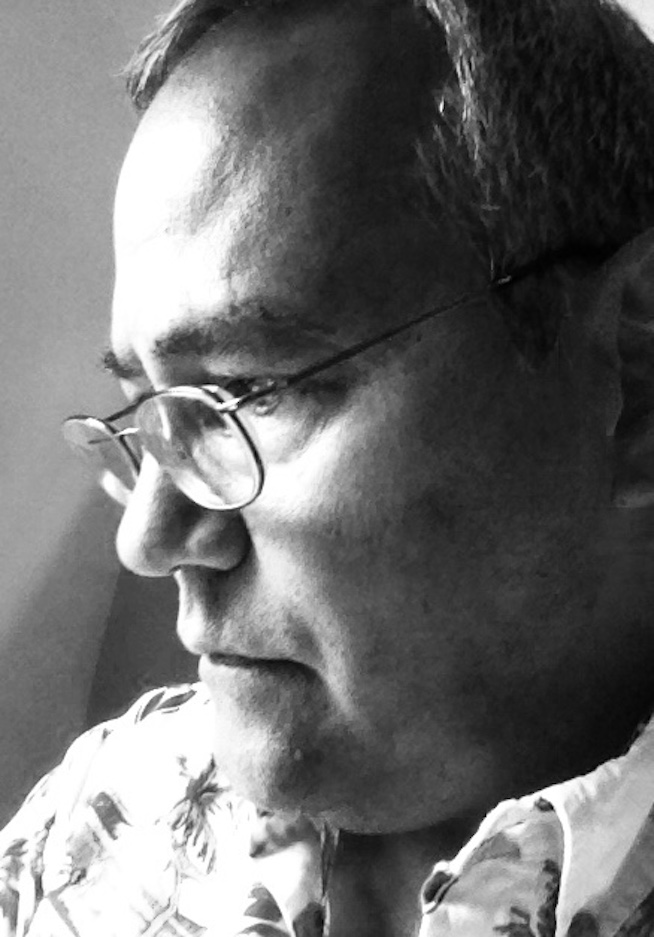 Black and white: A mixed-race white-Japanese man in contemplation by a sunny window. He wears a collared print shirt and a pair of glasses perched on his nose.