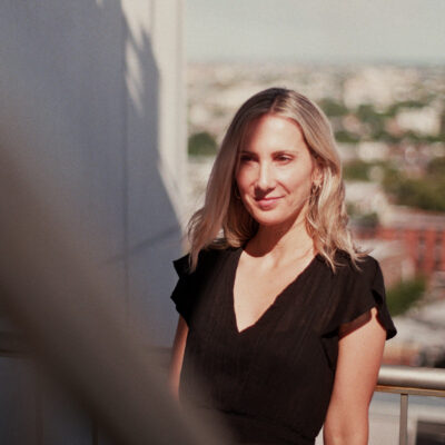 A Caucasian woman with a slight smile on her face stands in sunlight on the balcony of a tall apartment building. She has shoulder-length light blonde hair and wears a black v-neck top with short ruffled sleeves.