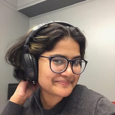 A young South Asian woman is sitting indoors and is turned to face the camera, smiling. She wears large black-framed glasses and a set of black headphones.