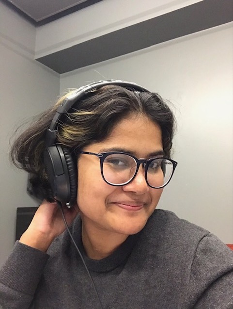 A young South Asian woman is sitting indoors and is turned to face the camera, smiling. She wears large black-framed glasses and a set of black headphones.