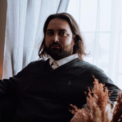 In this staged photo, Alejandro is seated in front of a tall window with white curtains. He is looking directly into the camera, and has brown eyes, dark eyebrows and beard, and shoulder- length brown hair. He is wearing a grey sweater over a white button-down shirt.