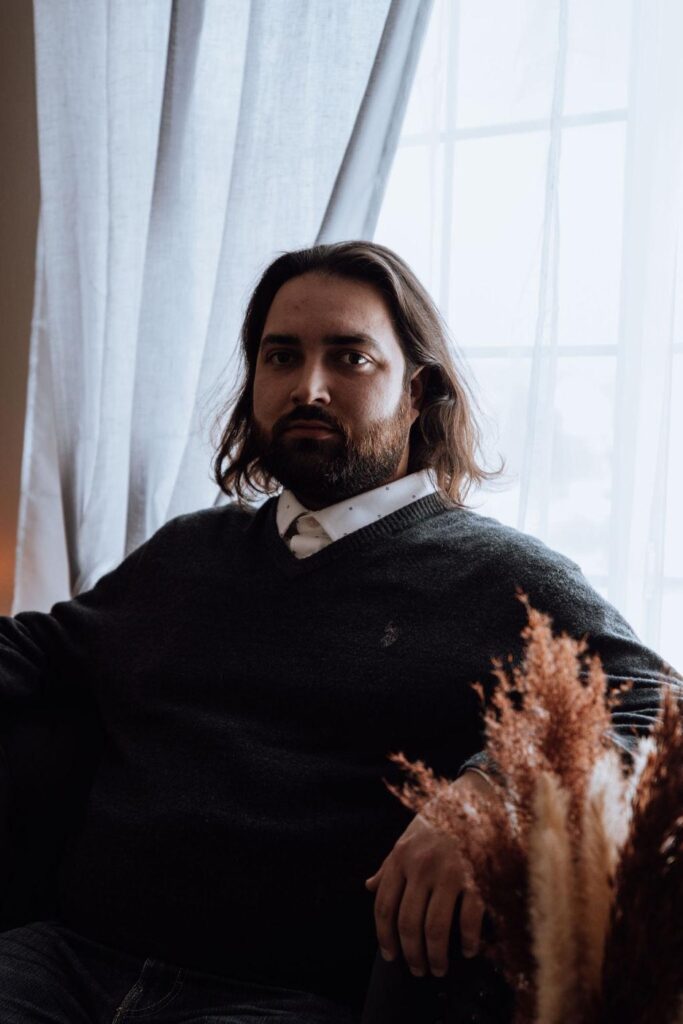 In this staged photo, Alejandro is seated in front of a tall window with white curtains. He is looking directly into the camera, and has brown eyes, dark eyebrows and beard, and shoulder- length brown hair. He is wearing a grey sweater over a white button-down shirt.