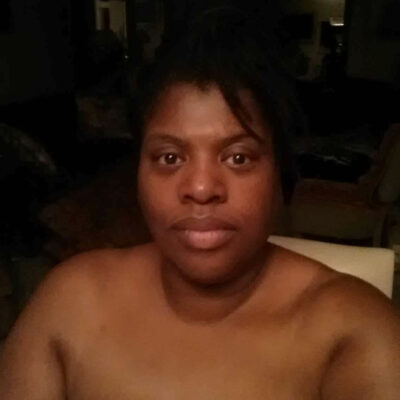 An African-American woman sits in a dark room. Her shoulders are bare.