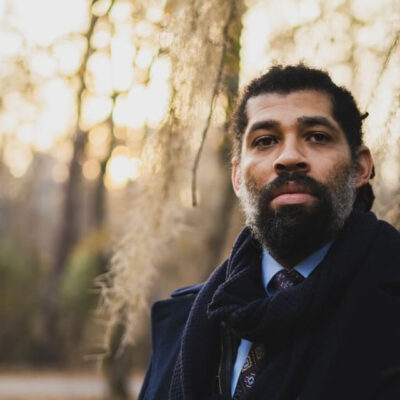 The poet, a Black man dressed in a tie, blue dress shirt, navy scarf, and navy wool coat, stands by a tree outdoors in forest light. He has a black beard and greying sideburns.