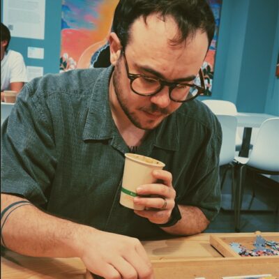 A white man with short brown hair, a beard and mustache, and glasses leans over a puzzle. He is wearing a short-sleeved dark cerulean button up and is holding a small coffee cup in his left hand. His right forearm is encircled by a tattoo of two slim black bands and his left wrist has a watch.