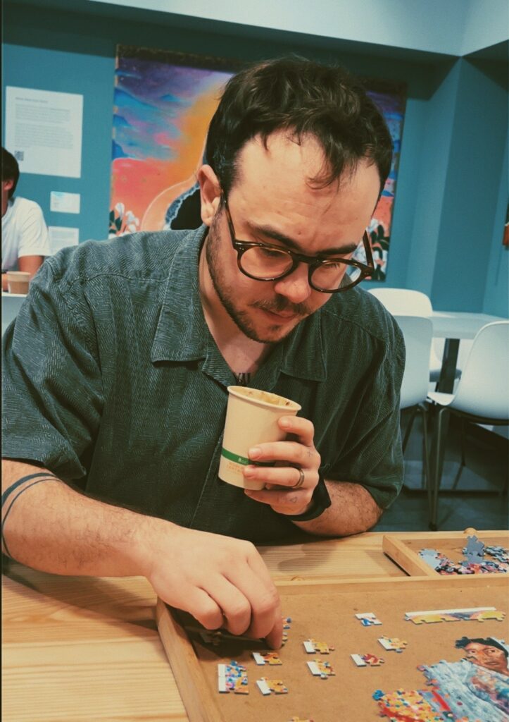 A white man with short brown hair, a beard and mustache, and glasses leans over a puzzle. He is wearing a short-sleeved dark cerulean button up and is holding a small coffee cup in his left hand. His right forearm is encircled by a tattoo of two slim black bands and his left wrist has a watch.