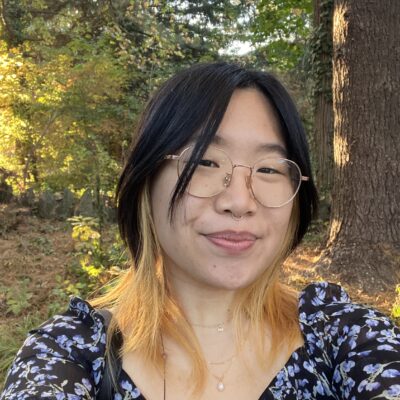 Monica Kim, a light-skinned Korean American woman with glasses and a nose ring, wearing a blue and black floral dress outdoors.