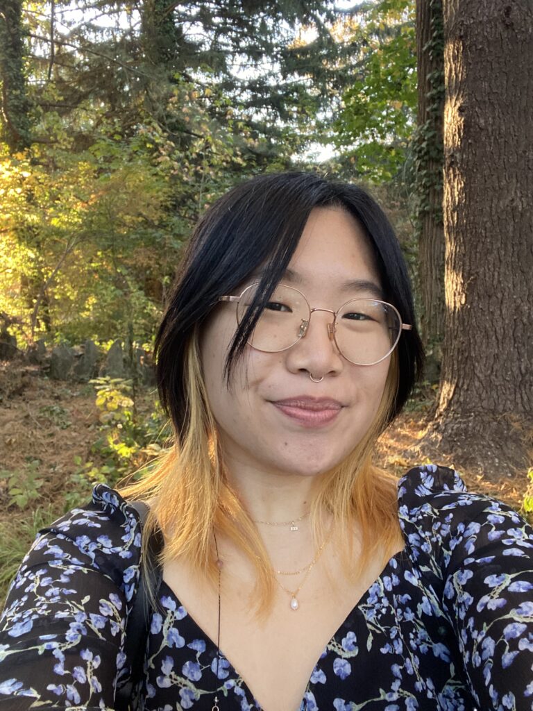 Monica Kim, a light-skinned Korean American woman with glasses and a nose ring, wearing a blue and black floral dress outdoors.