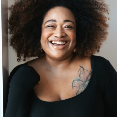 a fat black woman with a curly dark Afro smiles. She is wearing a black dress, and has a large flower tattoo on her upper left chest