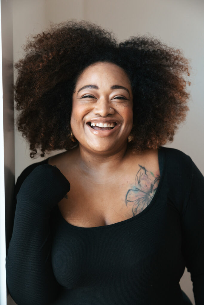 a fat black woman with a curly dark Afro smiles. She is wearing a black dress, and has a large flower tattoo on her upper left chest