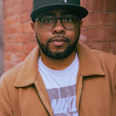 An African-American man stands in front of a red brick wall. He wears large black-framed glasses, a white Nike t-shirt, a camel-colored jacket, and a black cap that reads "Chi"