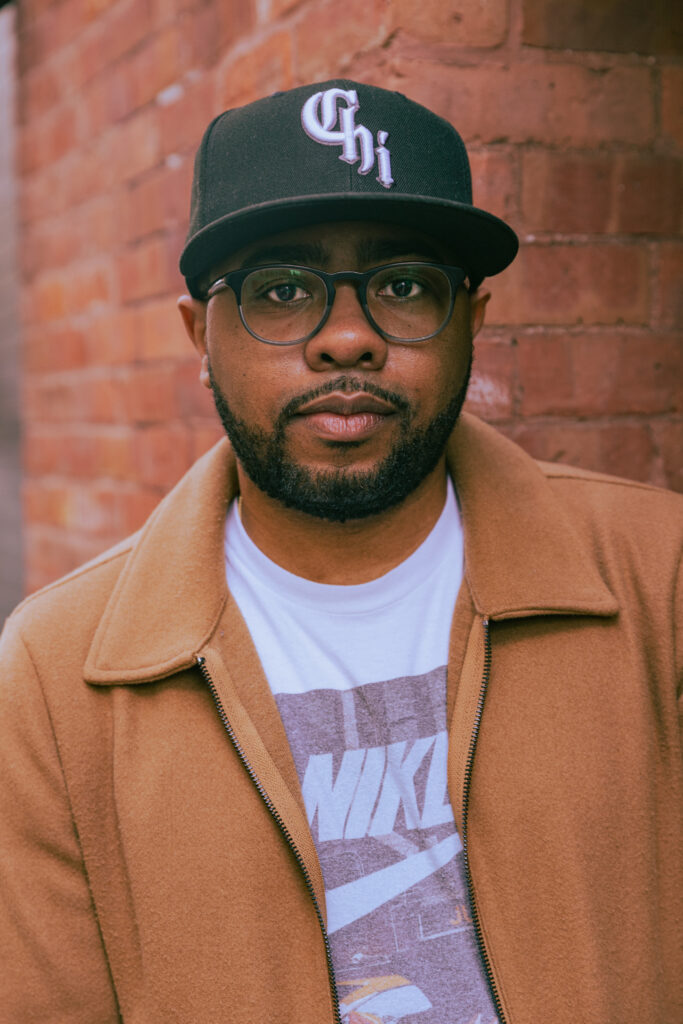 An African-American man stands in front of a red brick wall. He wears large black-framed glasses, a white Nike t-shirt, a camel-colored jacket, and a black cap that reads "Chi"