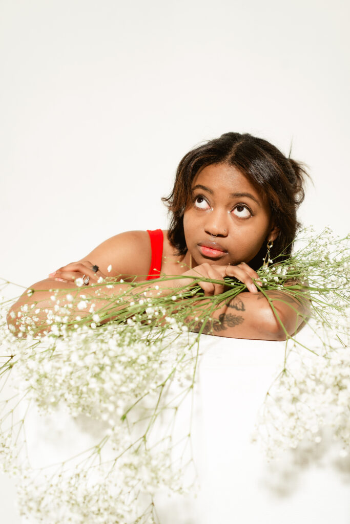 An African-American woman with a septum ring sits in a white well-lit room holding two bunches of small white flowers. Her eyes look upwards and she wears a red tank top.