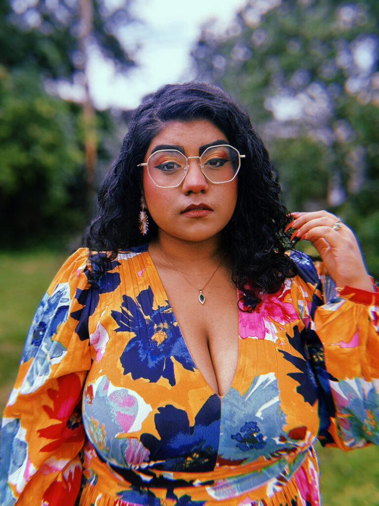 Head on photo of Indo-Guyanese woman with gold glasses in a yellow floral dress on a blurred green background.