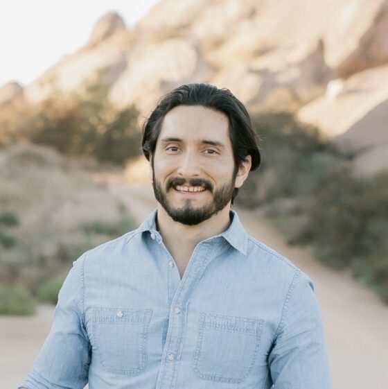 A dark-haired Latinx man with a beard and a chambray button-down shirt smiles as he stands outdoors.