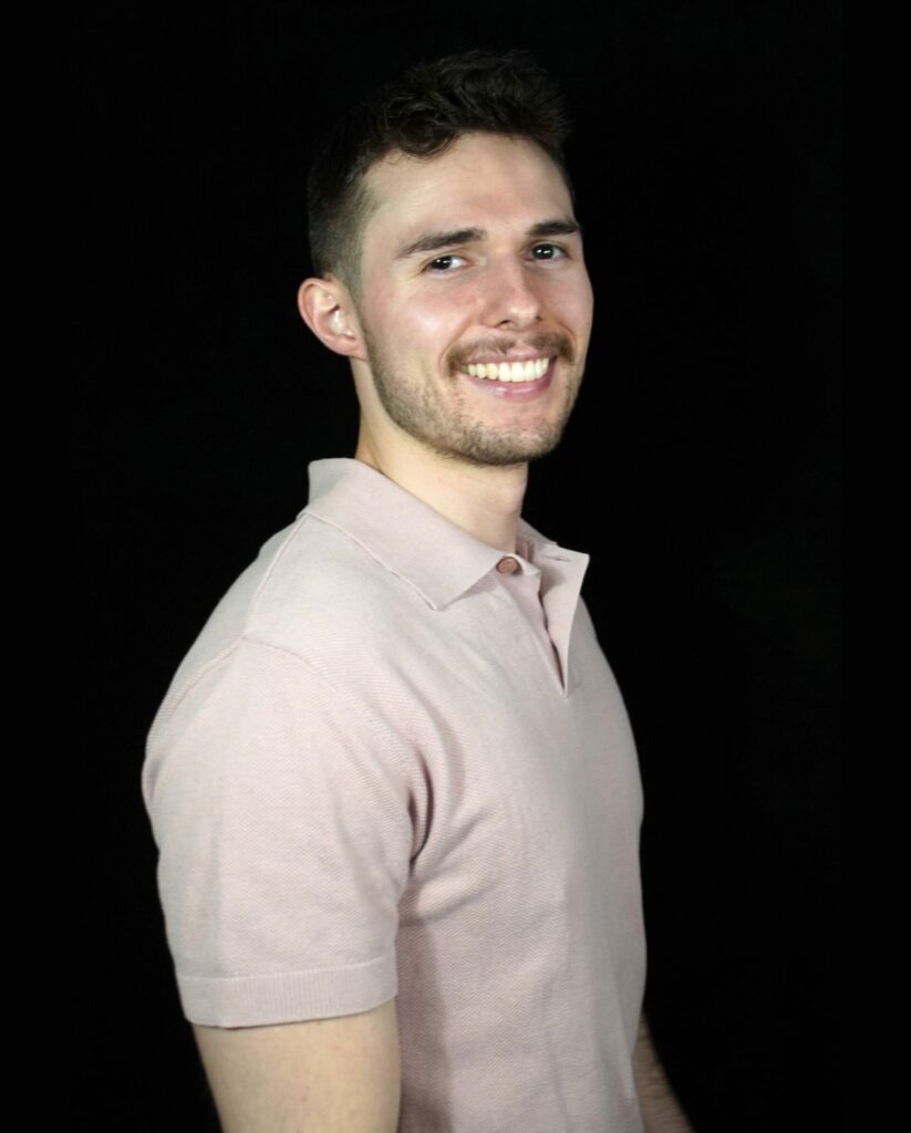 A young man with short dark hair and facial hair is smiling while posing against a black background. He is wearing a light pink short-sleeve polo shirt and is turned slightly to the side, looking at the camera.