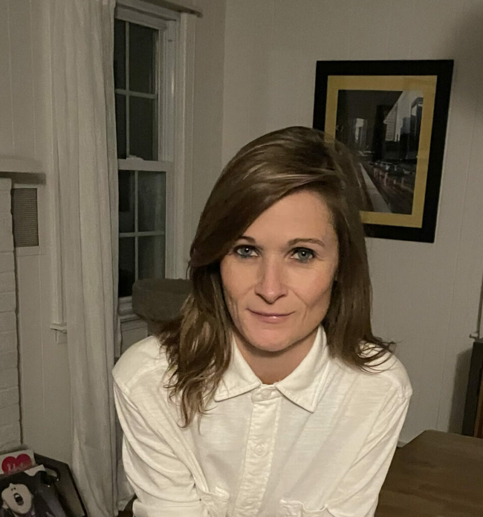 A woman leans over the corner of a table indoors. She has small smile on her face and wears a white button-down shirt, blue jeans, and black Chuck Taylors.