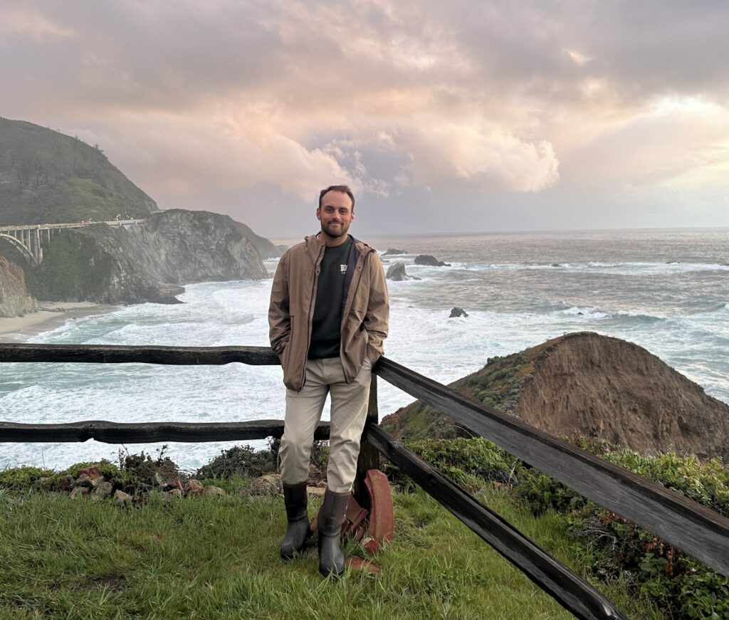 A man wearing a light brown rain jacket leans his back against a wooden railing at the edge of a grassy cliff. Behind him, the ocean is white with foam and a mountain rises, cut through by a bridge.