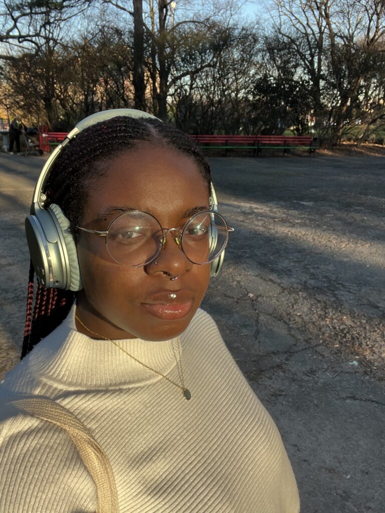 Photo of a person with dark brown skin and hair styled in cornrows. They are wearing a white turtleneck shirt, teal over-ear headphones, circular wire-framed glasses, and have a neutral expression on their face. A park bench and trees are visible in the background.