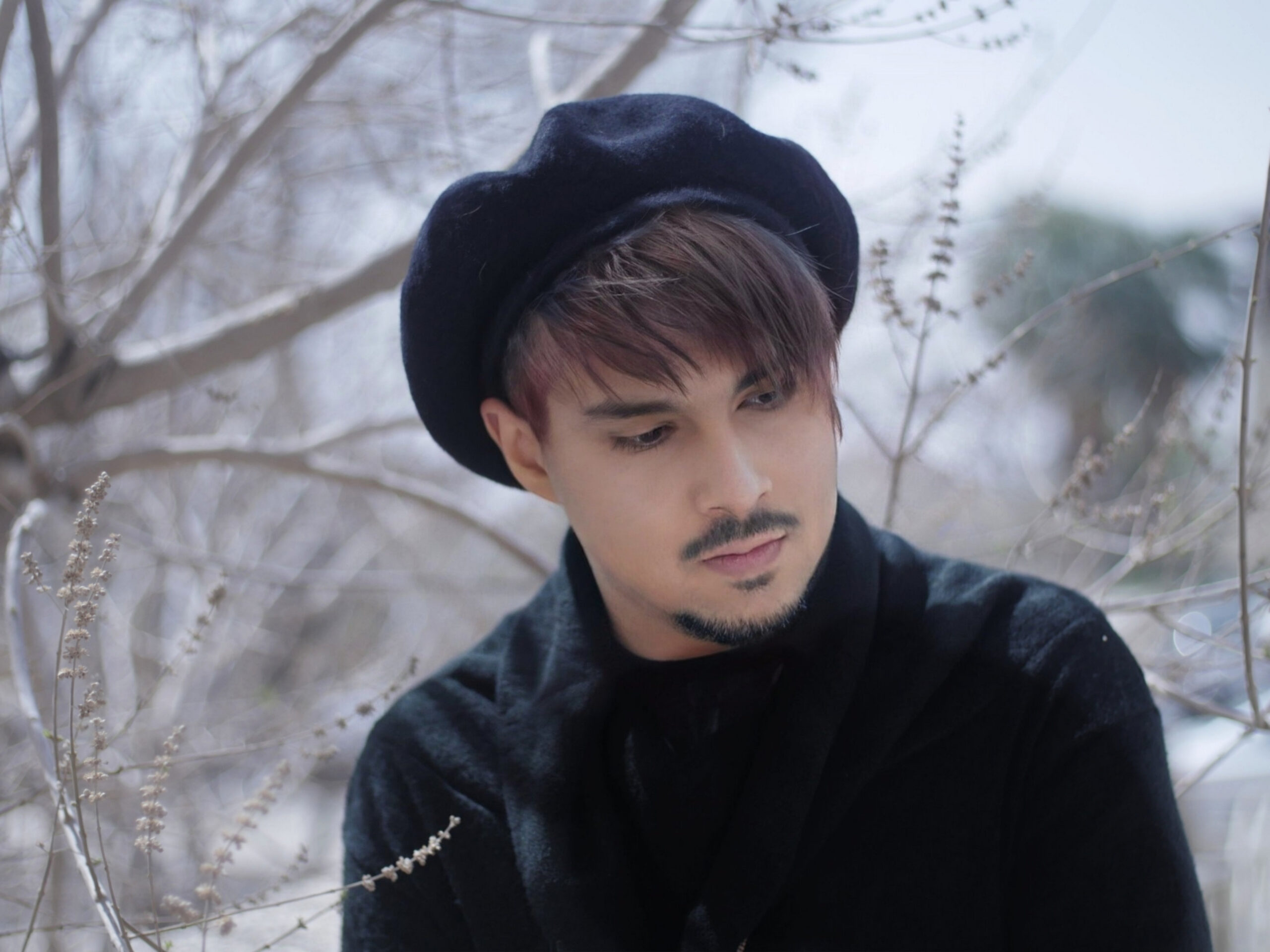 A person with short, reddish-brown hair & a neatly trimmed goatee wears a black beret & a black sweater, standing among leafless branches.