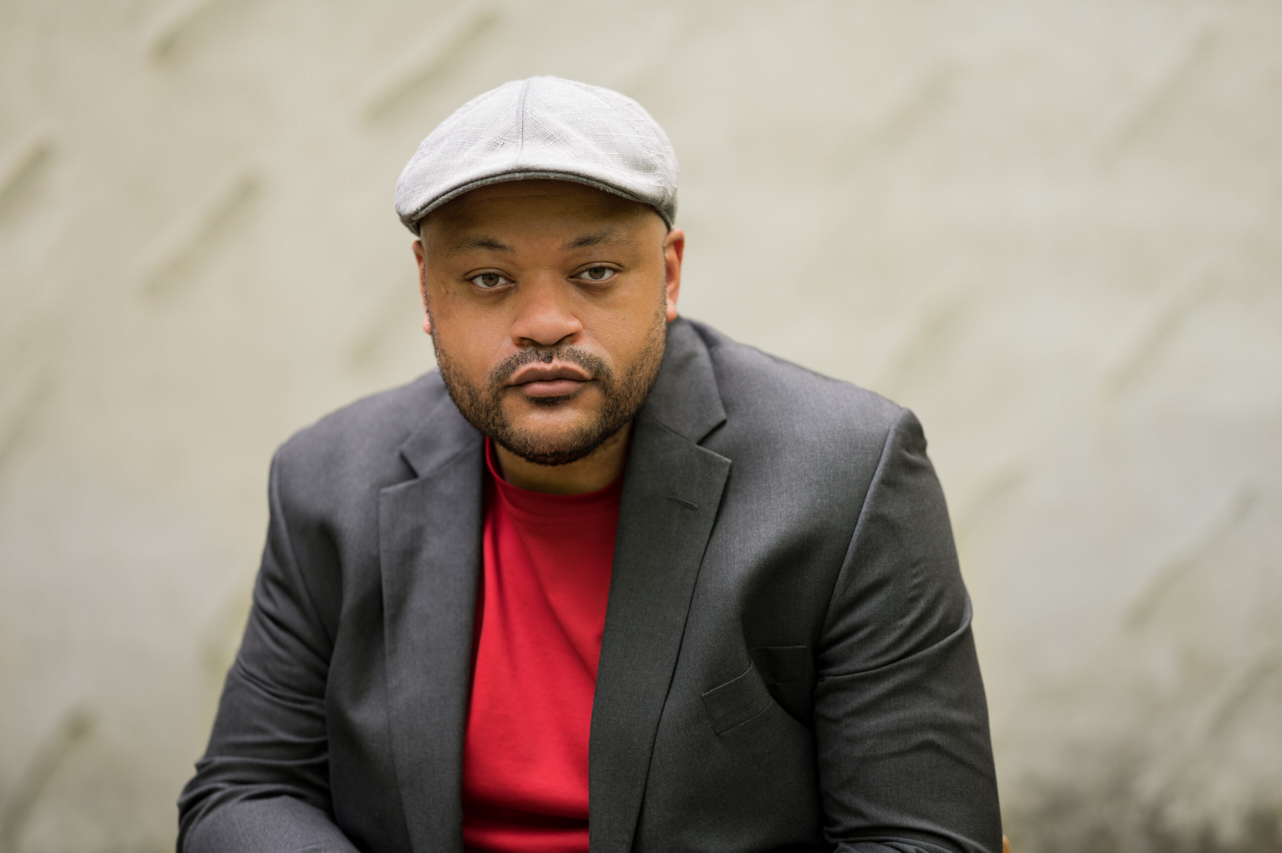 A photo of Christian J. Collier, a Black Poet, wearing a red t-shirt, grey blazer, and off-white flat cap. He is looking directly into the camera. 
