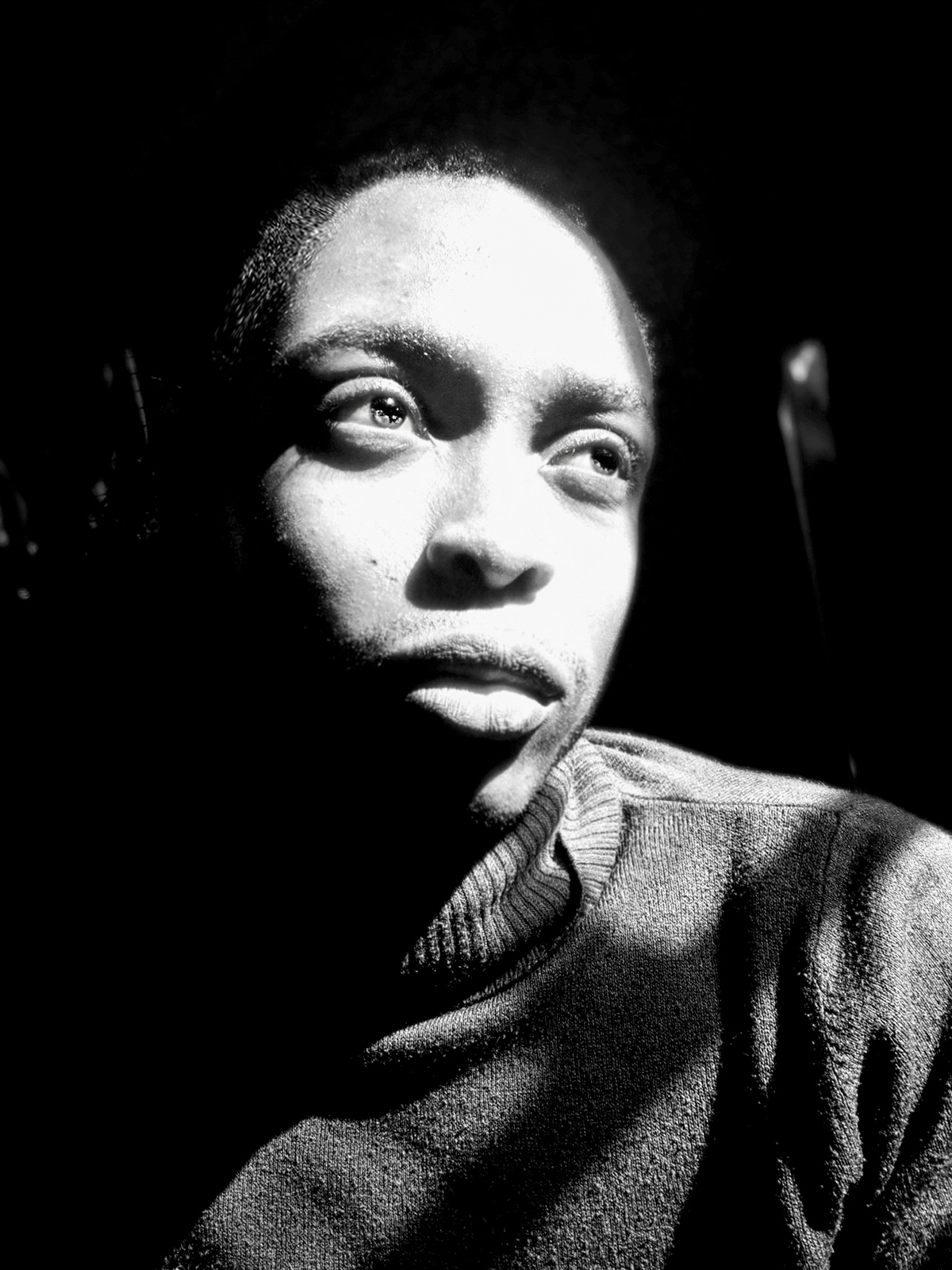 A black-and-white photo of a Black man gazing up to the right. His face is bathed in light, his gaze is soft, and he wears a turtleneck sweater.