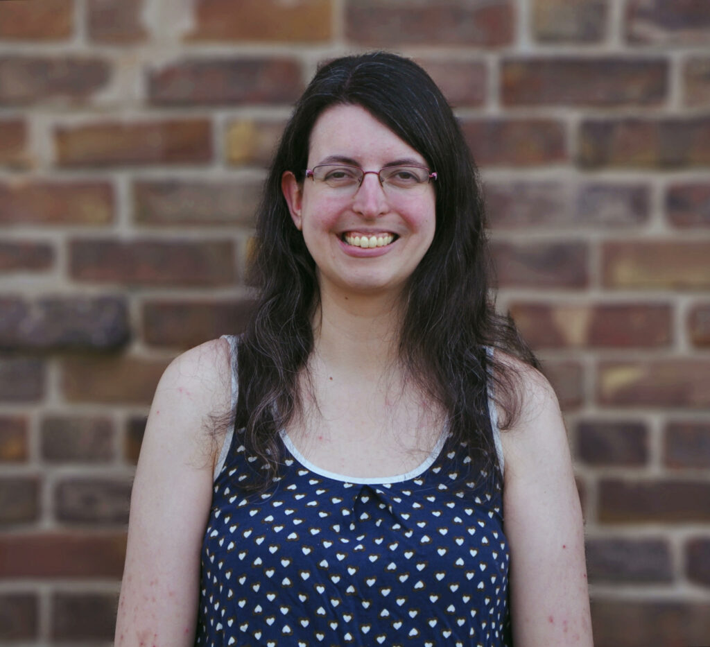 A fair-skinned woman with wavy dark brown hair that falls past her shoulders stands against a brick wall, smiling. She wears glasses and a scoop-neck sleeveless navy top patterned with small white hearts.