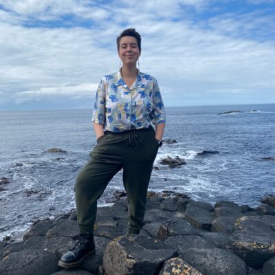 Jo Bear standing on the Giant’s Causeway in Northern Ireland. They have short dark hair and wear a short-sleeve button-down blue, yellow, cyan, and white print shirt, dark olive green sweatpants, Doc Marten boots, and dangling earrings. They have a nose ring and are standing with their foot on a rock, hands in pockets.