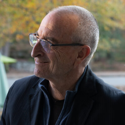 Photo of a man in profile wearing glasses and looking off-camera. He has a buzzcut and is wearing a navy blue collared jacket and a black shirt.