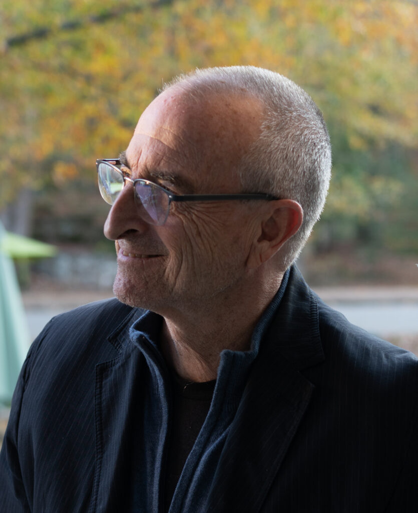 Photo of a man in profile wearing glasses and looking off-camera. He has a buzzcut and is wearing a navy blue collared jacket and a black shirt.