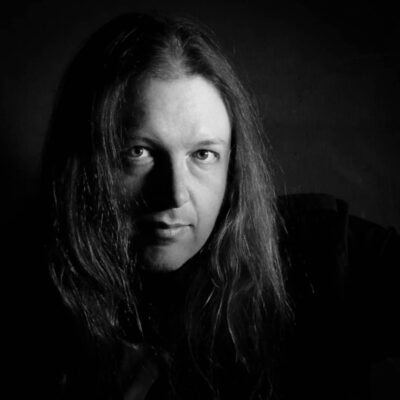Black-and-white photo of the poet, a Caucasian man with long hair parted at the center. His face is illuminated and he is looking at the camera with a neutral expression on his face.