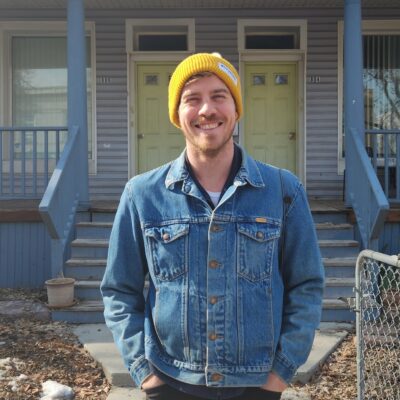 Poet Nicholas Pierce, a young Caucasian man wearing a yellow beanie and a denim jacket, smiles in front of a house with blue steps and yellow twin doors.