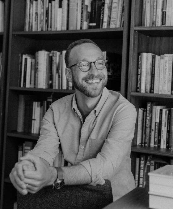 The poet Bobby Elliott, a Caucasian man with close-cropped light brown hair and a light brown heard, looks off to the side, smiling. His hands are clasped together in a relaxed pose and he wears brown round-rimmed glasses, a wristwatch, and a light blue button-down shirt. He sits in front of two large shelves of books.