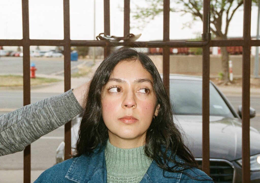 Jasmine Khaliq, a Pakistani Mexican poet, stands outside in front of a gate. She has brown eyes and long wavy black hair. She is wearing a light green turtleneck under a denim jacket and a small nose ring. Someone else's arm is extended behind her head and there is a grey car behind her and a parking lot and cargo train in the background.