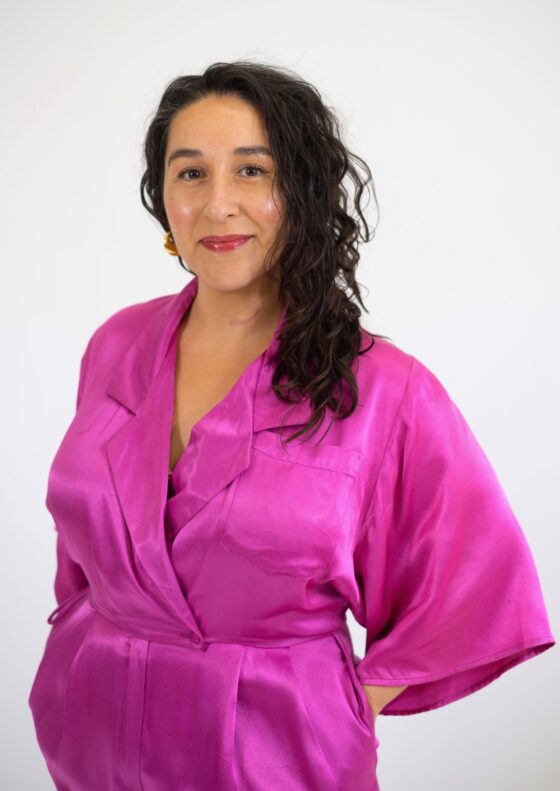 Jenna Martínez, a Mexican American poet, stands smiling in a silk pink robe. Her dark wavy hair is pulled to one side.