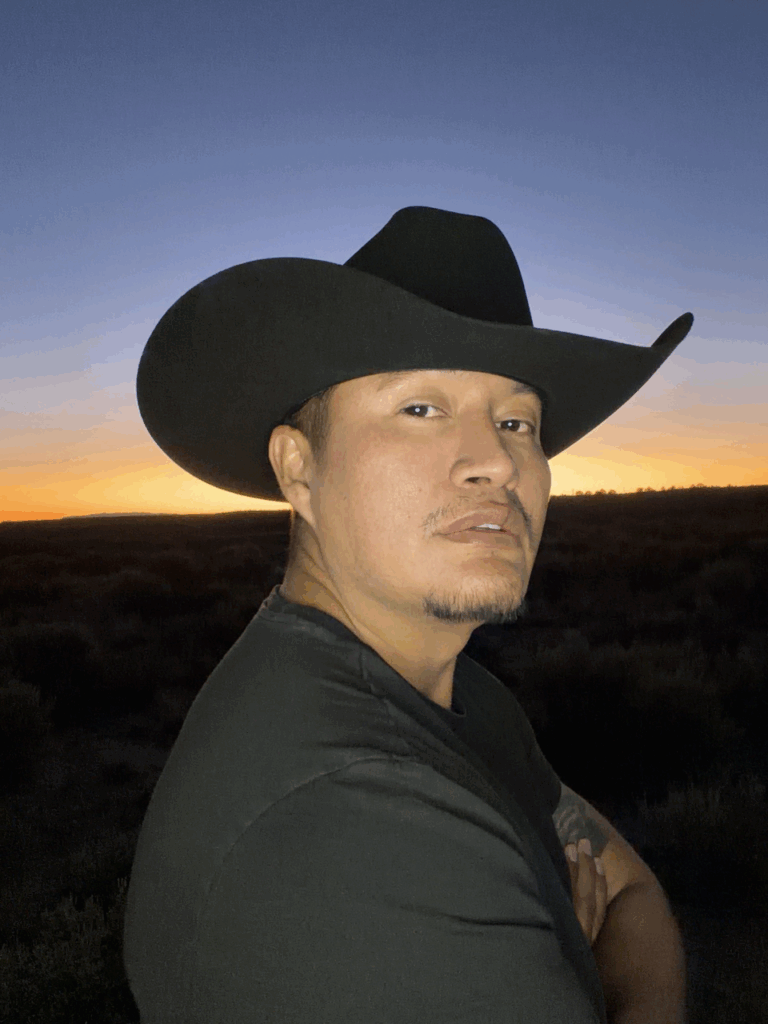 Diné poet J.K. Tsosie wears a black cowboy hat and a black tshirt. He stands outside in front of a setting sun. The sky is blue and orange.
