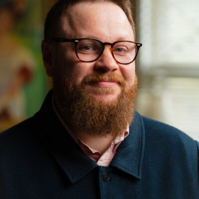 A man with short brown hair and a reddish-brown beard stands by a window indoors, smiling. He is wearing brown-rimmed glasses and a light pink collared shirt underneath a textured navy button-down shirt.